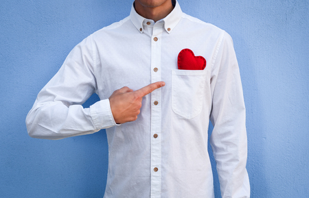 The guy in the white shirt points his finger at a red Valentine in his pocket against the background of the blue wall. Happy Valentine's day conceptの写真素材