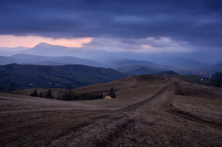 Camping with a tourist tent in a mountain village. Dusky autumn landscape. Stormy sky with scary clouds over a ridge. Unpaved country roadの写真素材