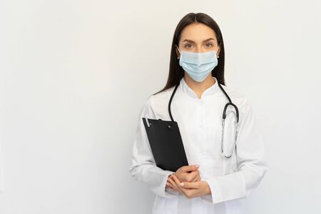 Friendly female doctor in a protective medical mask with a stethoscope, holds a folder in his hands,の写真素材
