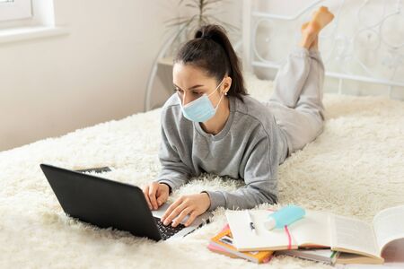 Young girl woman in protective medical mask on the bed, distance learning for students. Coronavirus. Quarantine. Woman look at a laptop computer. Freelancer, work at home, online education,の写真素材