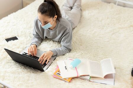 Young girl woman in protective medical mask on the bed, distance learning for students. Coronavirus....