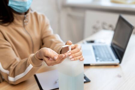 clouse up young girl woman wearing a protective medical mask distributes sanitizer to hands and then works with a laptop on the table during the COVID-19 quarantine. Coronavirus protection. Hygiene conceptの写真素材