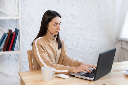 Young girl woman sits at a table, distance learning for students. Quarantine, self-isolation. Woman look at a laptop computer. Freelancer, distant work, work at home, online educationの写真素材