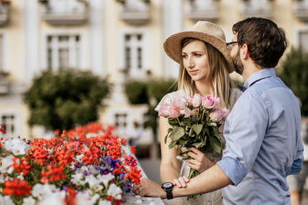 Close up of romantic couple are kissing in embrance with building on background. Girl in white dress with pink fresh flowers look awayの写真素材
