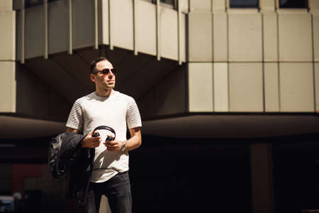 Portrait of smiling man with headphone in his hand, look away. Dj in black glasses and white T-shirt...
