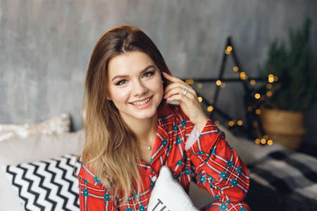 Close up of girl wears festive pajamas and take pillow in hands at home. Smiling woman in red night-wear on bedの写真素材