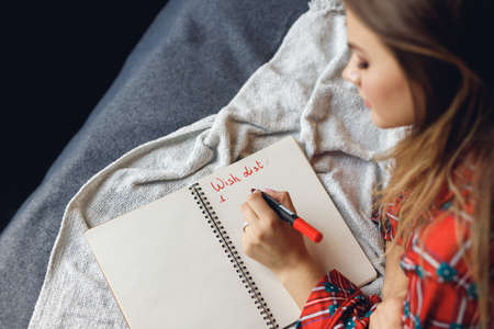 Close up of girl wears festive pajamas lies and write left hand at home. Woman in red night-wear on bed look down on focus Wish Listの写真素材