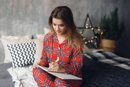 Girl wears festive pajamas and write wish list left hand at home. Serious woman in red night-wear on bedの写真素材