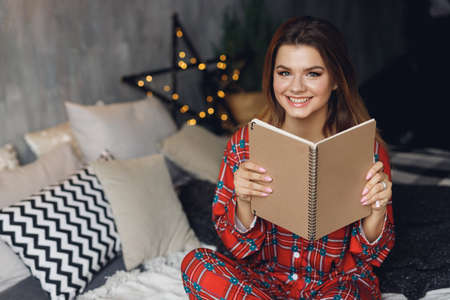 Close up of girl wears festive pajamas and take brown note in hands at home. Smiling woman in red night-wear on bedの写真素材