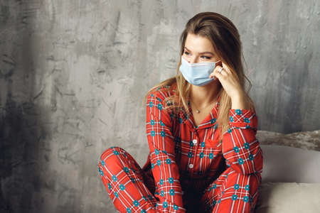 Close up of girl wears festive pajamas and medical mask at home. Cunning woman in red night-wear sit on bedの写真素材