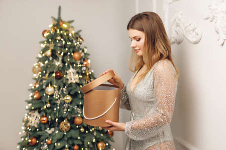 Close up of happy woman near festive Christmas Tree with gift. Concept of Christmas and New Year with girl in gray dress with gold boxの写真素材