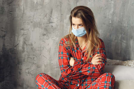 Close up of girl wears festive pajamas and medical mask at home. Offended woman in red night-wear sit on bedの写真素材