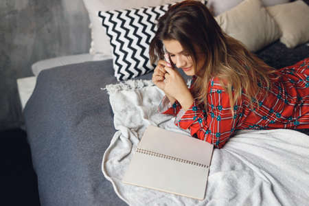 Top view of girl wears festive pajamas lies and write Wish List at home. Smiling woman in red night-wear on bedの写真素材