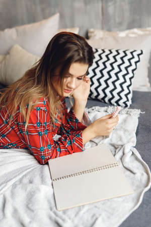 Close up of girl wears festive pajamas lies and write Wish List at home. Smiling woman in red night-wear on bedの写真素材