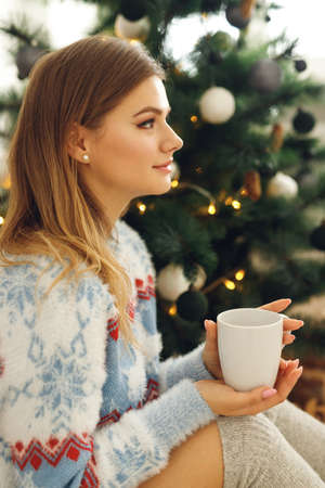 Close up of girl wears festive clothes take cup of coffee at home near Christmas Tree. Dreamy woman sit on mat in blue sweater with deer and gray stockingsの写真素材