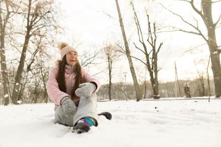 Photo of smiling young woman sitting on snow. Close up of happy girl wear gray gloves and pink hat outdoorの写真素材