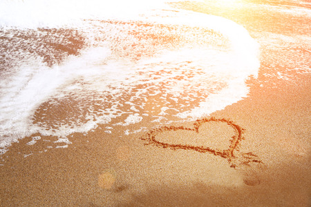 Soft wave of blue ocean on sandy beach. Heart drawn on the sand of the beach with sea foam and wave. Romantic vacation.の写真素材