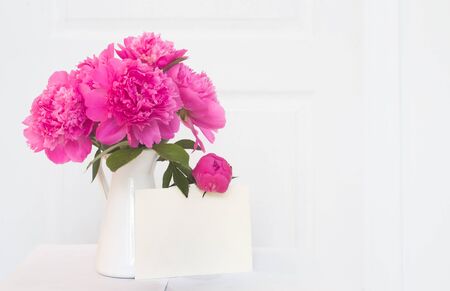 Pink peonies in white enamelled vase. Beautiful flowers in interior design. White paper for invitation text, white peonies in a vase, interior decoration.の写真素材