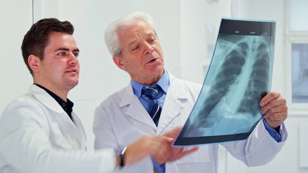Two caucasian male doctors looking at x-ray at the hospital. Senior gray man in white coat holding x-ray in his hand. Young brunette male medic showing something on x-ray to his elder colleagueの写真素材