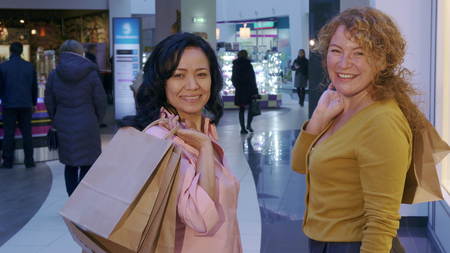 Two women turning back to the camera at the mall. Caucasian and asian ladies finishing their shopping. Two attractive female shoppers smiling for the camera with shopping bags on their shouldersの写真素材