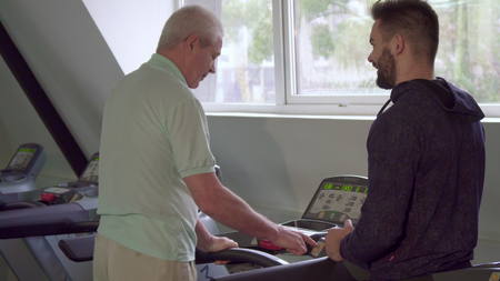 Senior gray man turning on the treadmill at the gym. Young bearded trainer advicing his aged client...