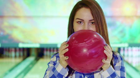 Pretty caucasian girl looking out from behind the bowling ball. Close up of young brunette woman holding red bowl in her hands. Attractive female bowler hiding her face behind red sphereの写真素材