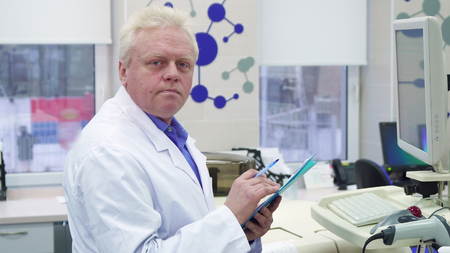 Senior male scientist noting information on his cliboard. Gray mature man in white coat writing on...
