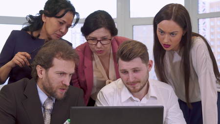 Business people looking at the laptop at the office. Two caucasian male managers sitting at the table. Three businesswomen standing behind two bearded manの写真素材