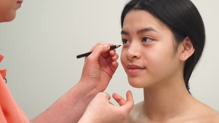 Beauty specialist using black cosmetic pencil to make marks on womans face. Pretty asian girl smiling while cosmetologist drawing lines under her eye. Attractive brunette client going to make plastic surgery at the cosmetology centerの写真素材
