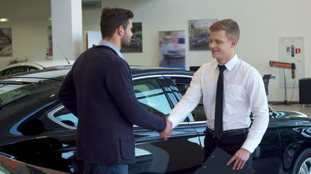 Attractive blond salesman showing something on clipboard to his client. Young male sales manager dealing with brunette bearded man at the dealership. Two caucasian handsome guys shaking hands against background of black carの写真素材