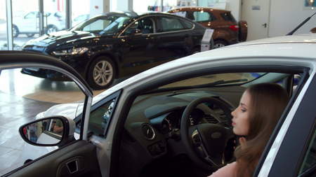 Pretty young woman sitting into the car at the dealership. Close up of attractive caucasian girl...