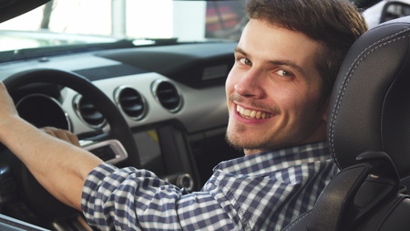 Close up shot of a young male driver using his smart phone sitting in a new convertible, smiling happily to the camera. Attractive young man enjoying his newly bought convertible auto.の写真素材