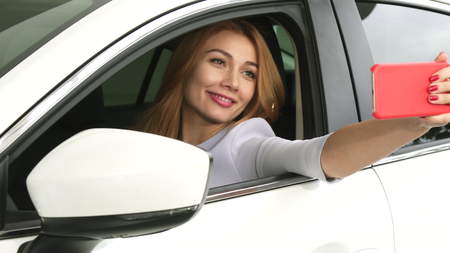 Cheerful young beautiful long haired woman sitting in a new car using smart phone taking selfies smiling joyfully emotions happiness driving travelling consumerism technology socialの写真素材