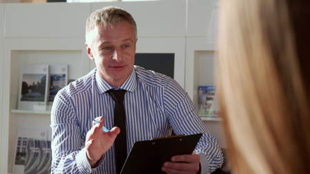 Salesman explaining contract conditions to the client at the car dealership. Close up of mature gray man holding pen and clipboard in his hands. Middle aged sales manager dressed in striped shirt and black tie talking with customerの写真素材