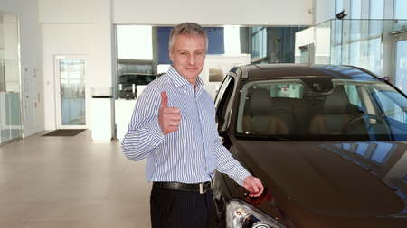 Attractive mature man showing his thumb up near the car at the dealership. Middle aged gray client approving new black SUV. Caucasian male client holding his hand on the car hoodの写真素材