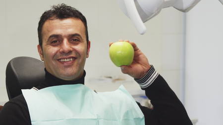 Handsome cheerful mature Hispanic male patient sitting in a dental chair smiling holding delicious green apple health happiness teeth smile dentistry medicine food consciousness.の写真素材