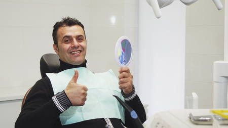 Cheerful handsome mature male patient sitting in the dental chair looking in the mirror smiling showing thumbs up curing cavity treatment health medicine dentistry clinical examination.の写真素材