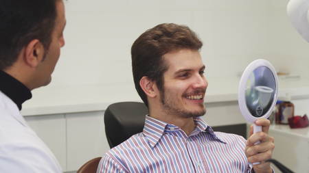 Close up of a cheerful handsome young man smiling looking in the mirror after dental procedure talking to his dentist occupation profession client patient medicine happiness smile teeth dentistry.の写真素材