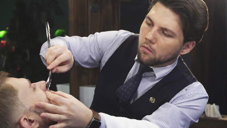Professional barber working at his barbershop shaving his young male client with a razorの写真素材