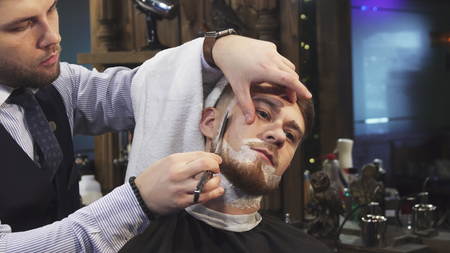 Close up of a handsome young man getting his beard shaved at the barbershop professional barber using a razorの写真素材