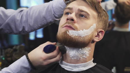 Handsome young man getting his beard shaved at the barbershop professional barber applying shaving cream on his face and neckの写真素材