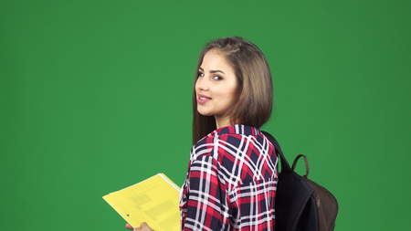 Rear view shot of a young beautiful happy female student with books and a backpack smiling joyfully to the camera over her shoulder on green backgroundの写真素材