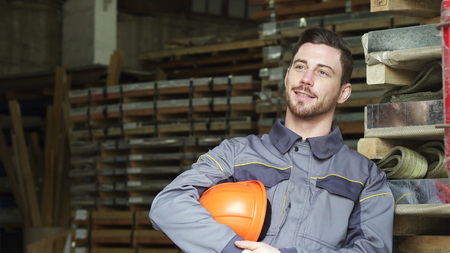 Handsome cheerful young industry worker smiling looking away taking off his hardhat resting after working at the factory warehouse metalworking profession success busy positive concept.の写真素材
