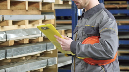 Cropped close up of a metalworking factory warehouse worker in protective uniform holding his hardhat checking inventory in stock using his clipboard walking away storage business logistics.の写真素材