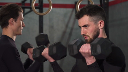 Close up shot of a bearded handsome sportsman concentrating exercising with dumbbells at the gym. Two sportsmen lifting weights training together at crossfit box. Effort, power, masculinity.の写真素材