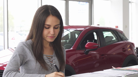 Attractive woman buying a new car at the dealership and signing papers for insurance. Young female customer filling documents for her new automobile. Consumerism, ownership concept.の写真素材