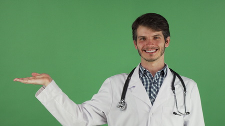 Portrait of a young handsome cheerful male doctor smiling happily holding copy space on his hand. Attractive friendly practitioner posing on green chromakey background.の写真素材