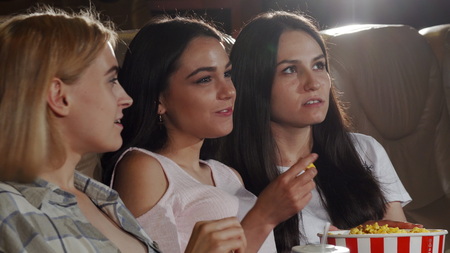 Young beautiful female friends enjoying eating popcorn and watching a movie together at the local cinema. Happy female friends smiling to the camera at the movie theatre.の写真素材