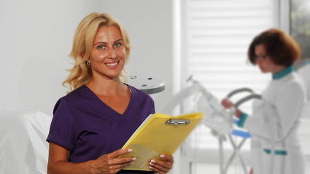 Mature beautiful female therapist checking medical papers while her colleague operating hospital equipment on the background. Attractive doctor smiling to the camera while working.の写真素材