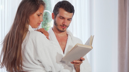 Happy couple wearing bathrobes reading a book together. Handsome man hugging his gorgeous girlfriend while reading her a book. Loving young couple resting at home, enjoying reading near the window. Relationships, lifestyle concept.の写真素材